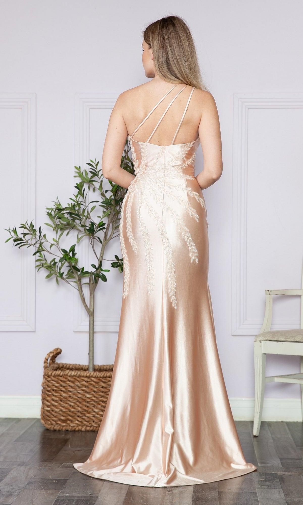 A woman wearing the One-Shoulder Embellished Long Prom Dress 9358 stands with her back turned in a room with a potted plant, white chair, and light walls.